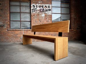 Diese Sitz-Bank ist für einen großen Esstisch. Die Bank ist aus massiver Eiche gefertigt. Die Seiten der Bank sind auf Gehrung mit der Sitzfläche verbunden. Die Holzmaserung wird von der Sitzfläche nahtlos an den Seiten weitergeführt. Die Rückenlehne ist mit schwarz pulverbeschichteten Winkeln aus 8mm Stahl stabil mit der Bank verbunden. Eine Strebe unter der Sitzfläche sorgt für zusätzliche Stabilität. Das schlichte Design überzeugt durch die klare Linienführung. Die großen Holzflächen mit ausgesuchtem Holzbild geben der Bank ein warmes und einladendes Aussehen.